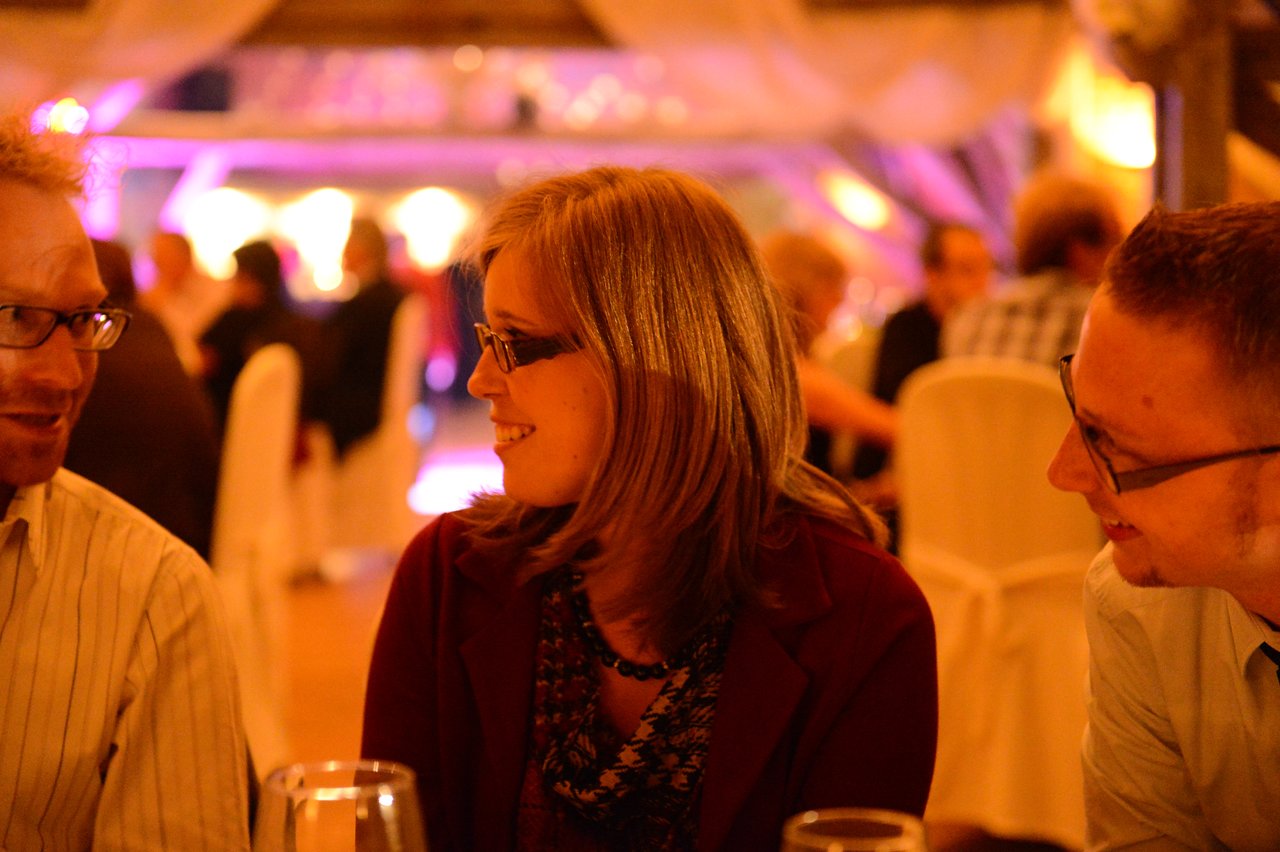 A woman in glasses smiles while talking to two men at a wedding reception.