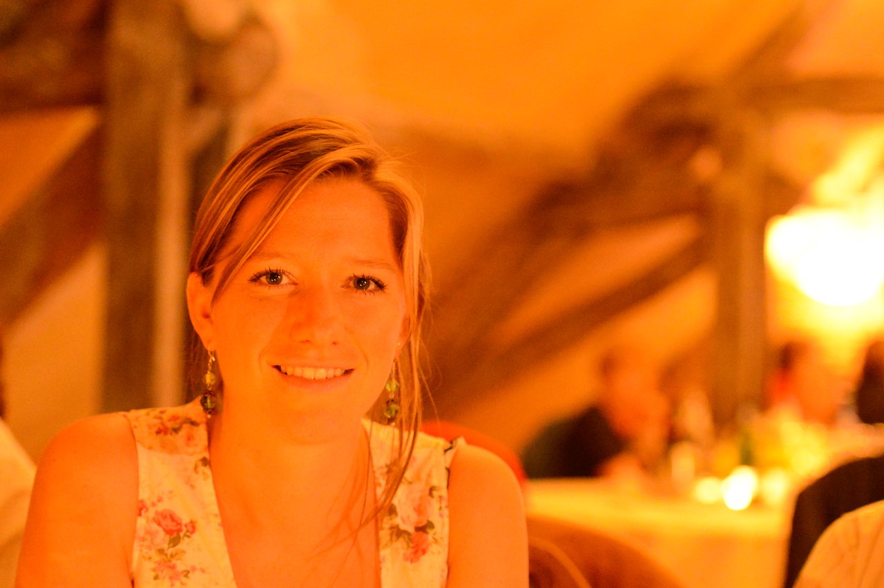 A woman smiles at the camera in a warmly lit wedding reception setting with guests in the background.