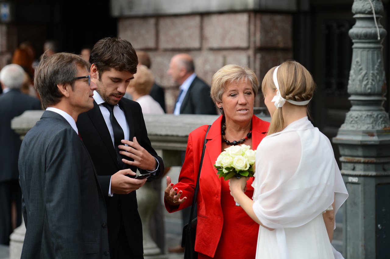 A bride holding flowers talks with three well-dressed guests outside a building during a wedding gathering.