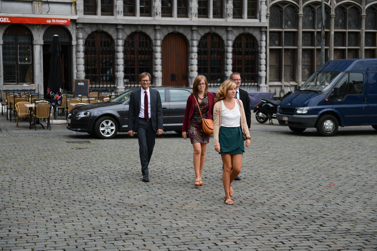 Four people in formal and semi-formal attire walk across a cobblestone square, possibly arriving for a wedding event.