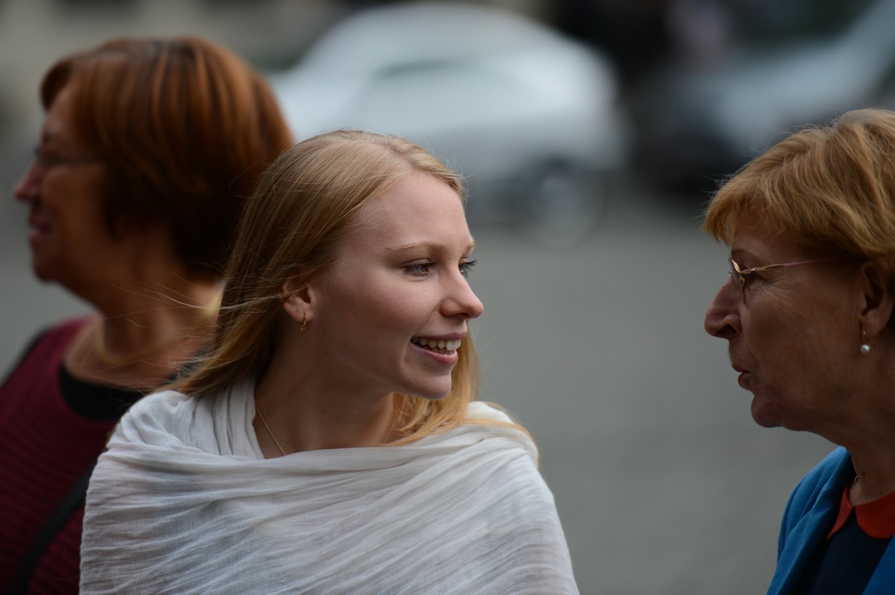 A woman in a white shawl smiles while talking to an older woman wearing glasses.