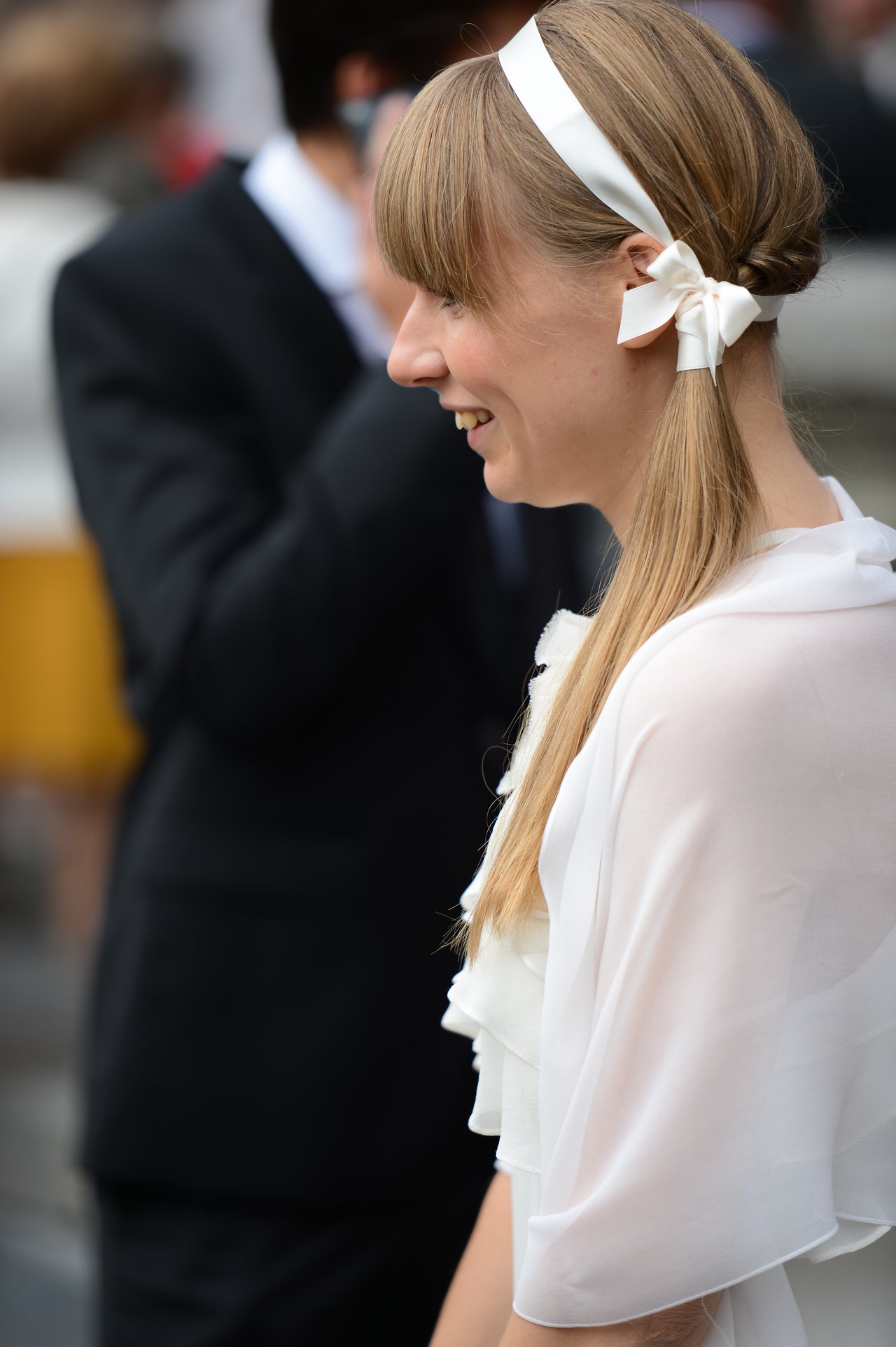 A smiling woman in a white outfit with a ribbon in her hair at a wedding celebration.