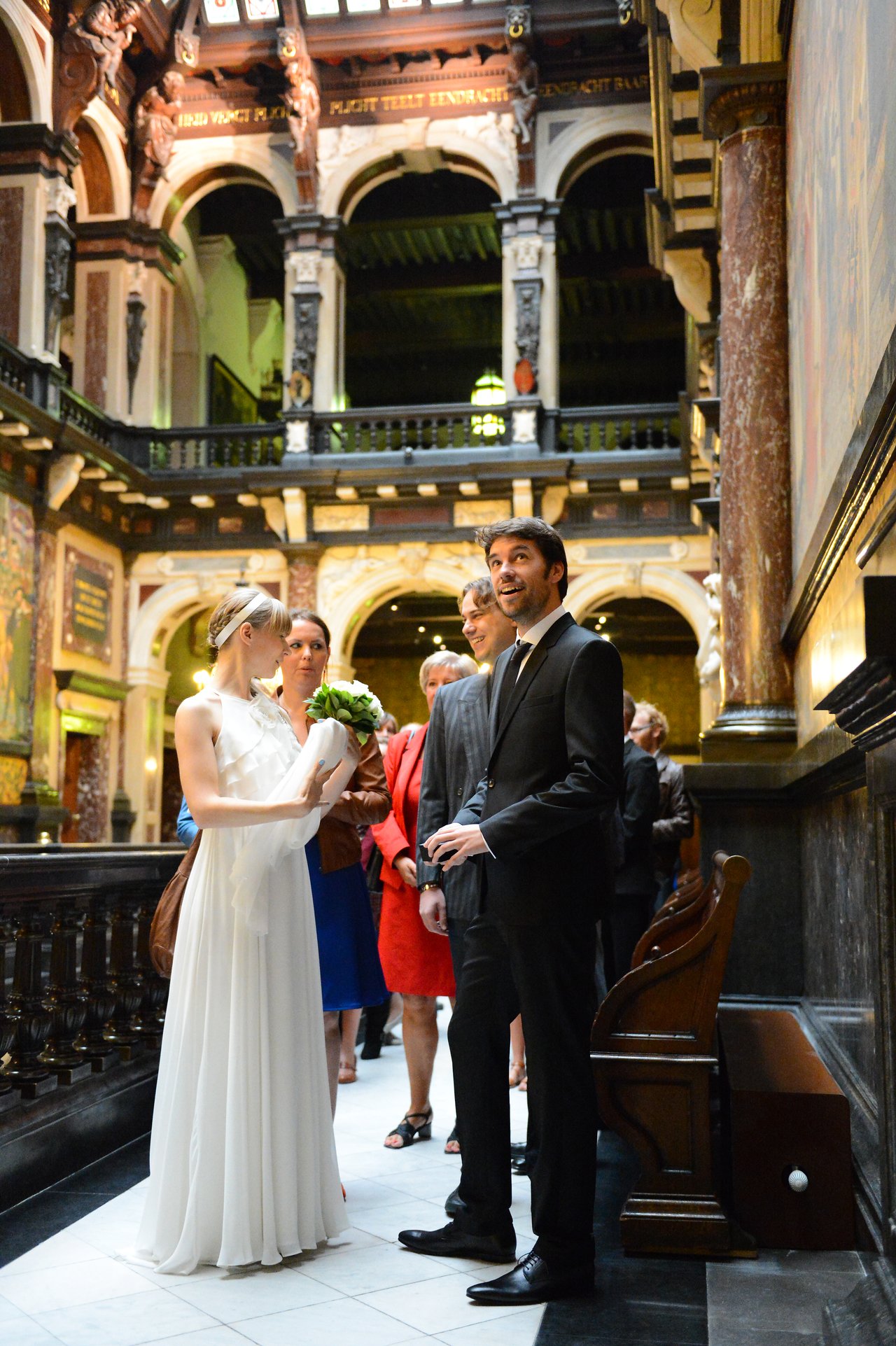 A bride in a white dress holds a bouquet while talking to the groom in a suit, surrounded by guests.