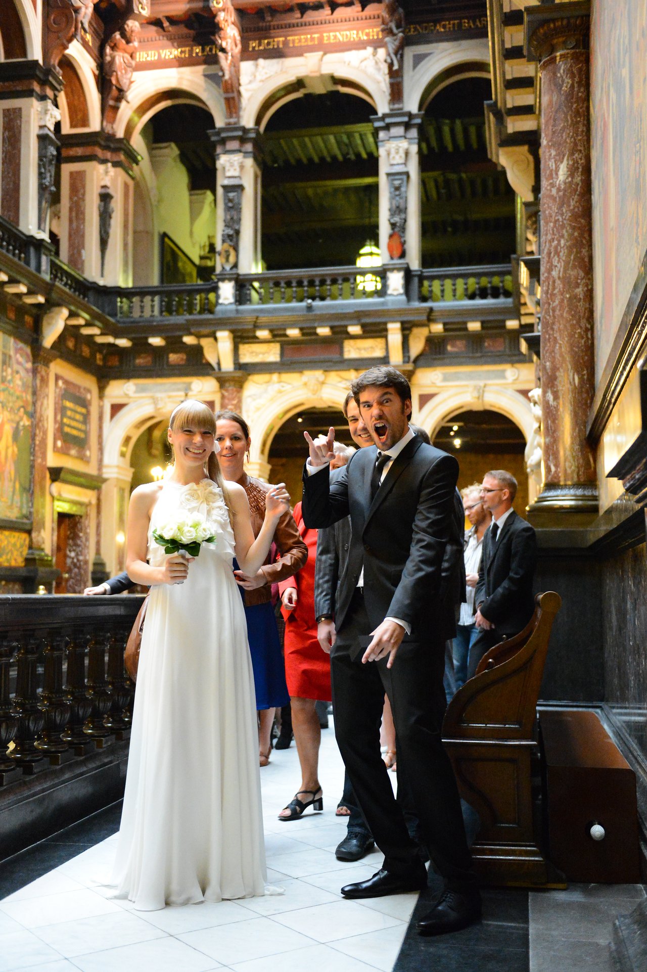 A bride in a white dress holds flowers while the groom playfully gestures, surrounded by wedding guests in an ornate hall.