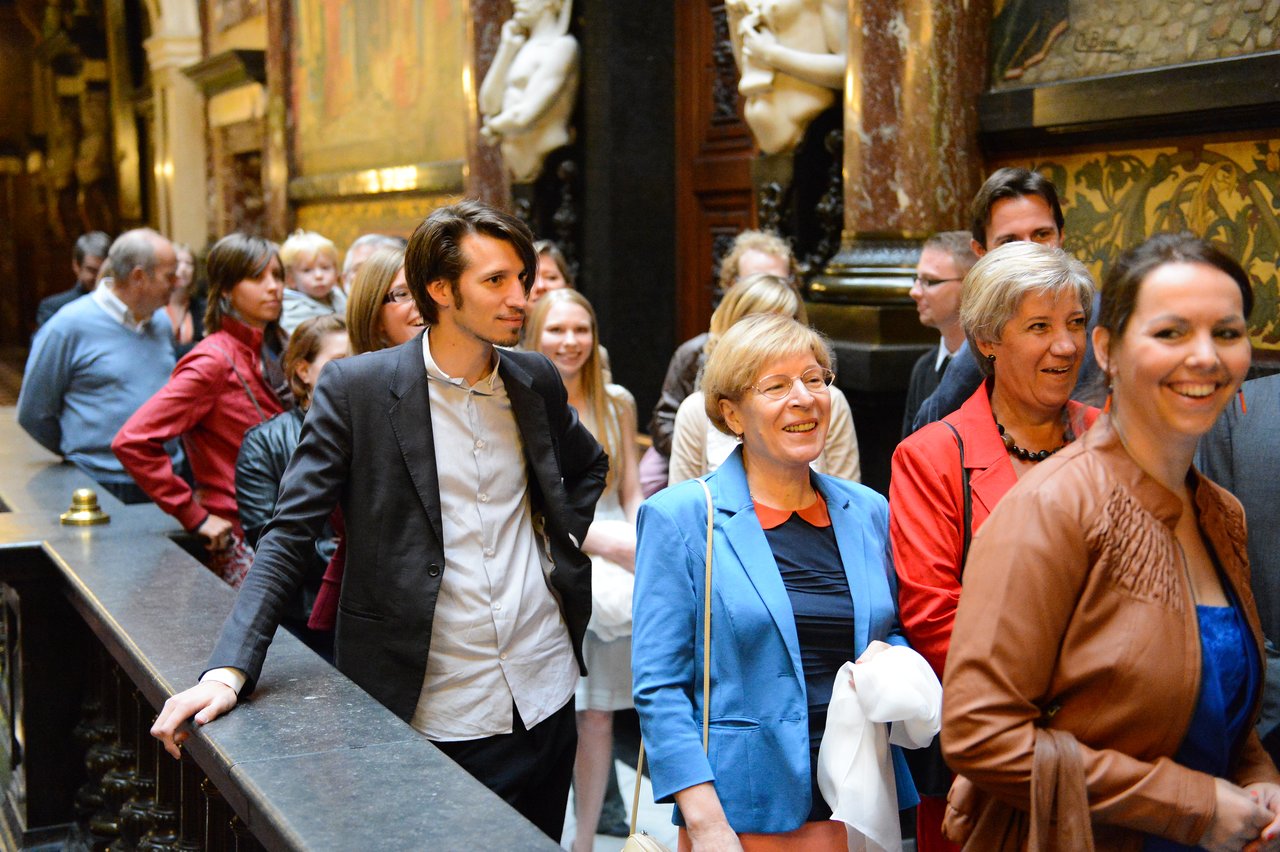 A group of wedding guests smiles and walks through an ornate indoor venue, dressed in formal and semi-formal attire.