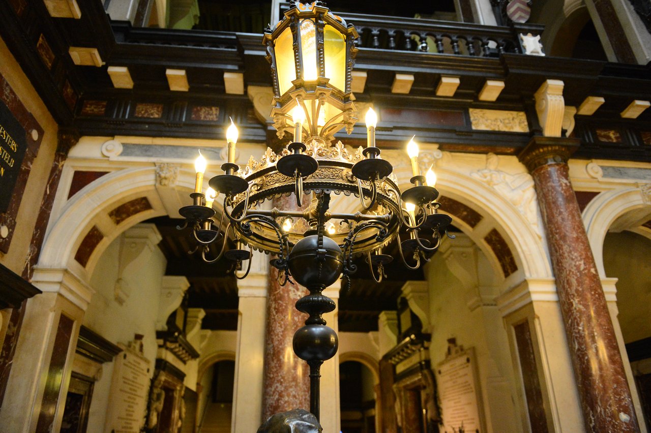 An ornate chandelier with lit candles hangs in a historic building with decorative arches and columns.
