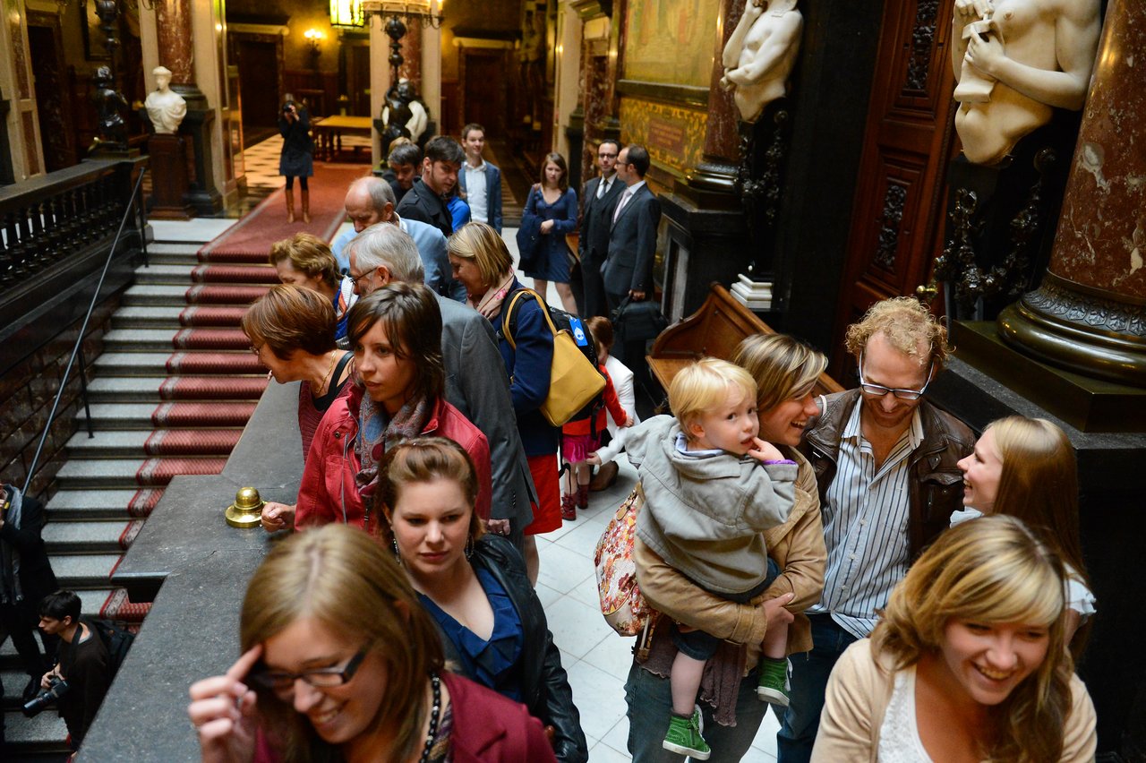 A group of wedding guests walks through an ornate hallway, chatting and smiling, with a child being carried.