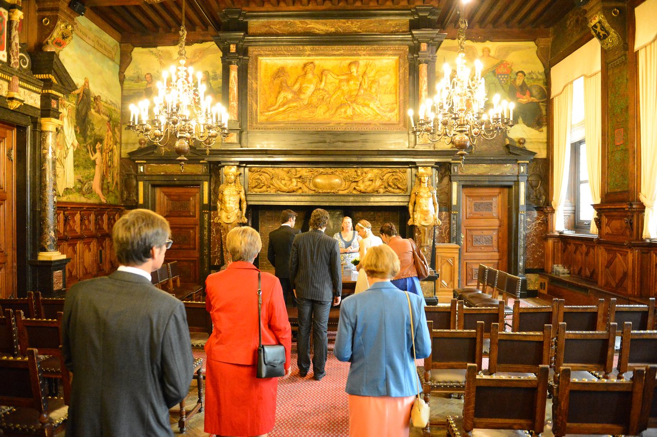A wedding ceremony in an ornate room, with guests walking toward the couple and officiant at the front.