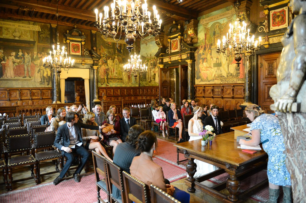 A bride and groom sit at a table while a woman officiates, with guests seated behind them.
