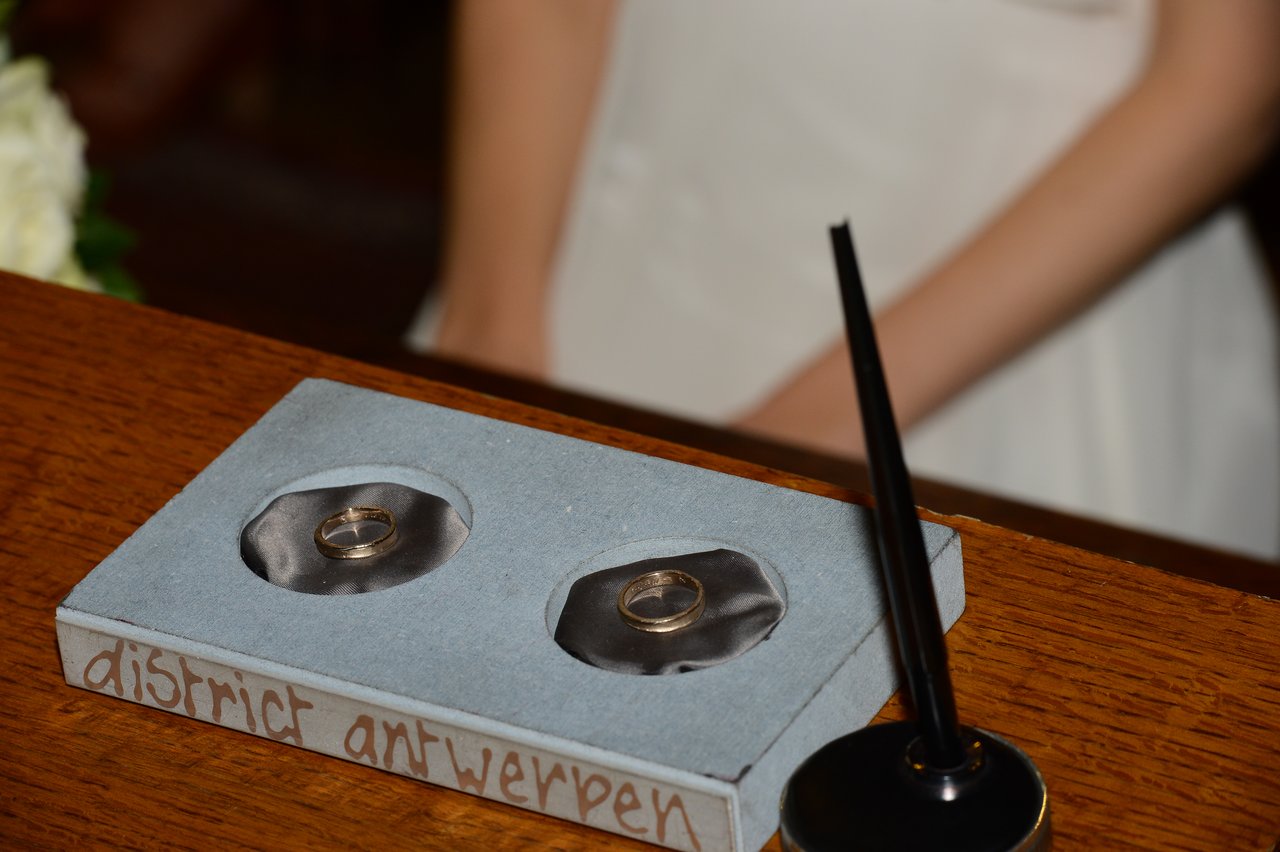 Two wedding rings rest on a small tray, with a bride in a white dress standing nearby.