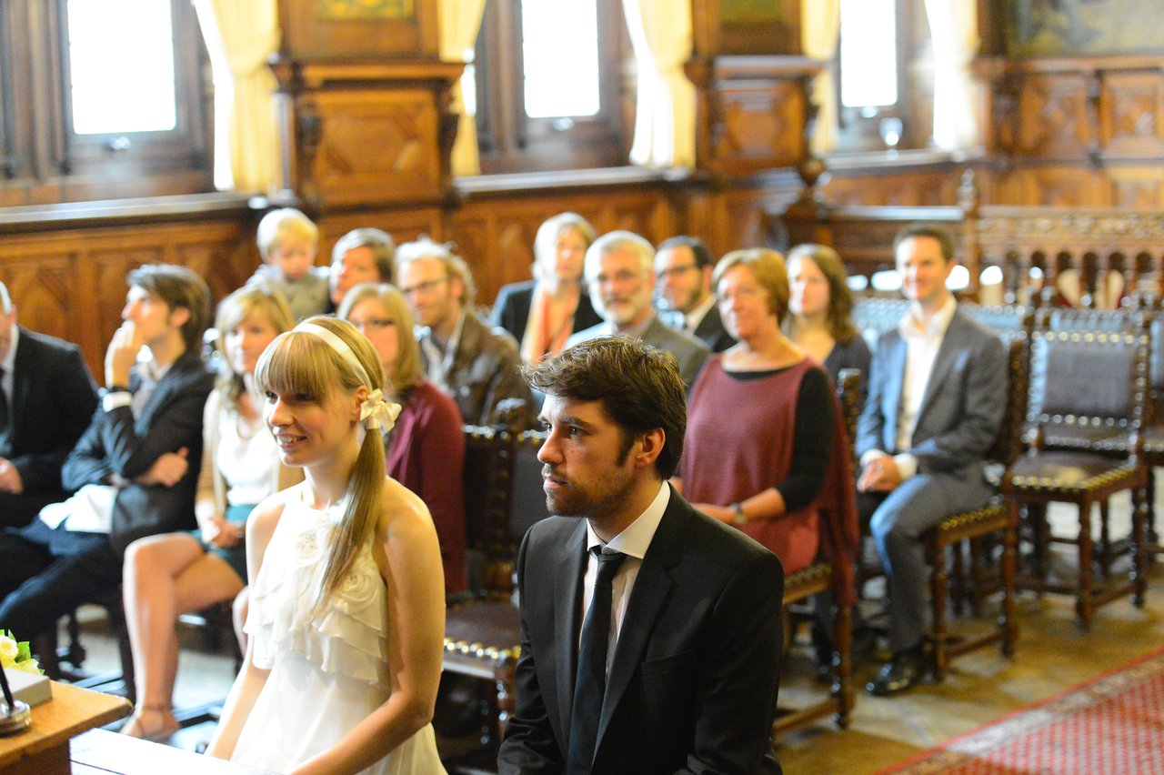 A bride and groom sit at the front of a room during their wedding ceremony, with guests behind them.