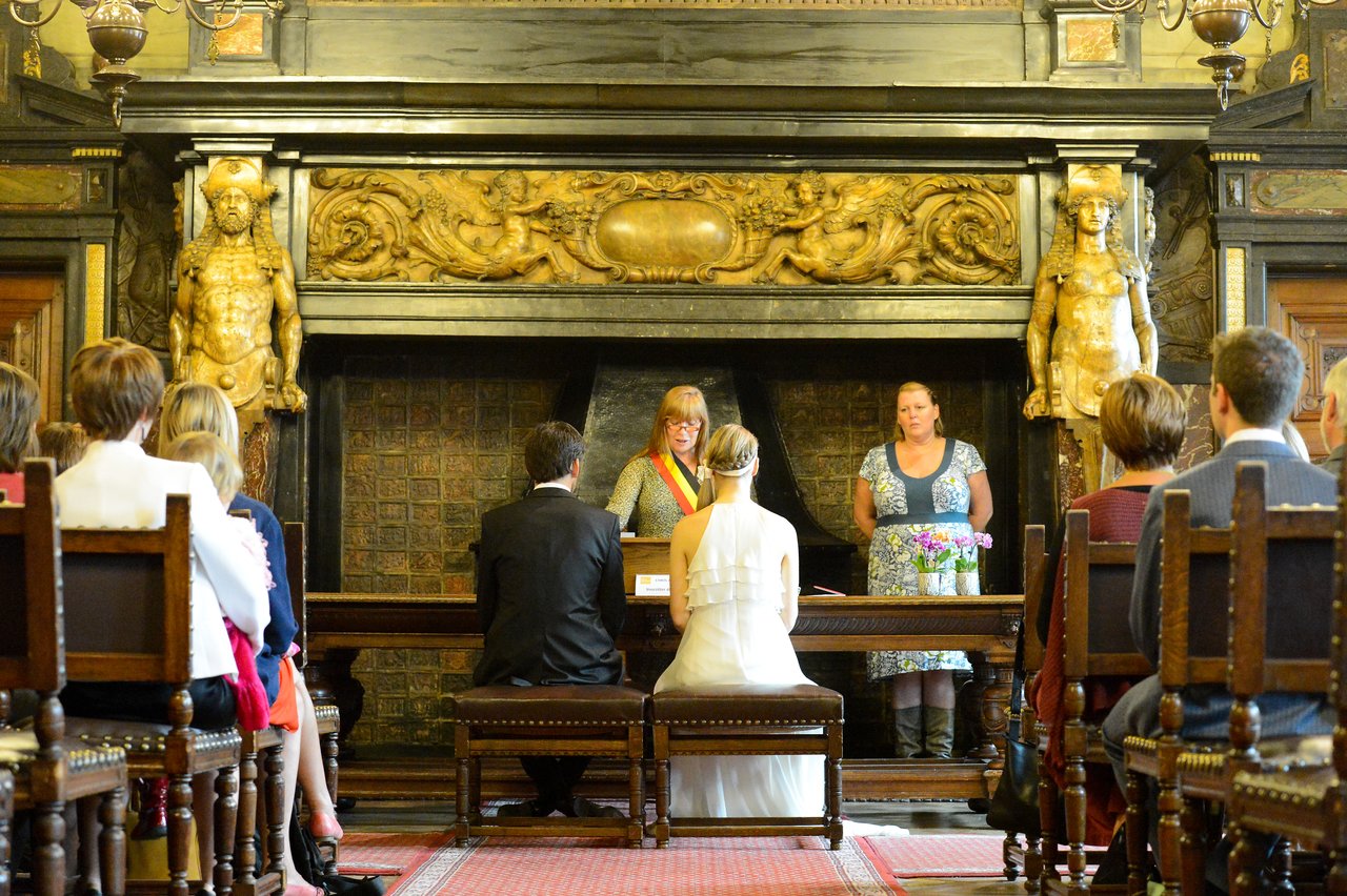 A bride and groom sit before an officiant during their wedding ceremony, surrounded by seated guests in a historic room.