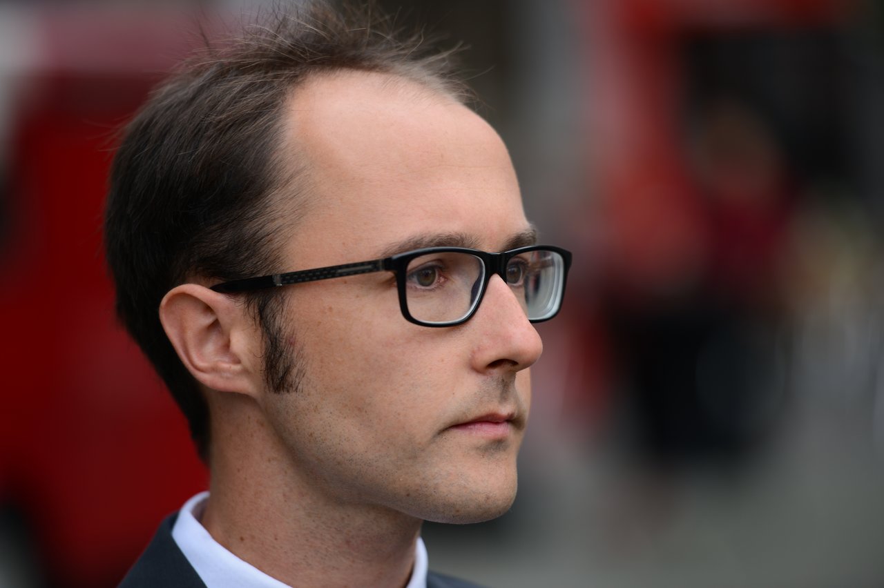 A man in glasses and a suit looks ahead with a serious expression during a wedding event.