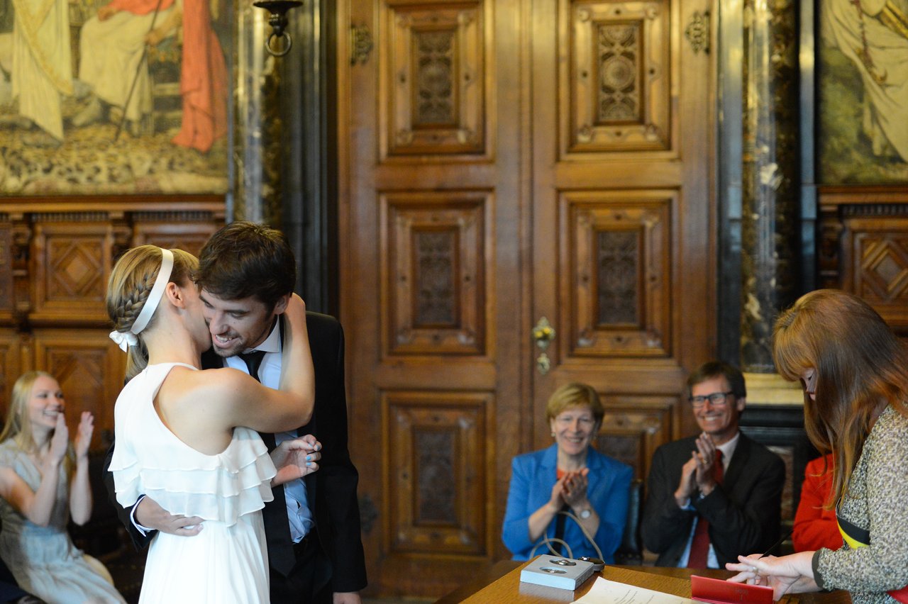 A newlywed couple embraces and smiles during their wedding ceremony, while guests clap and a registrar officiates.