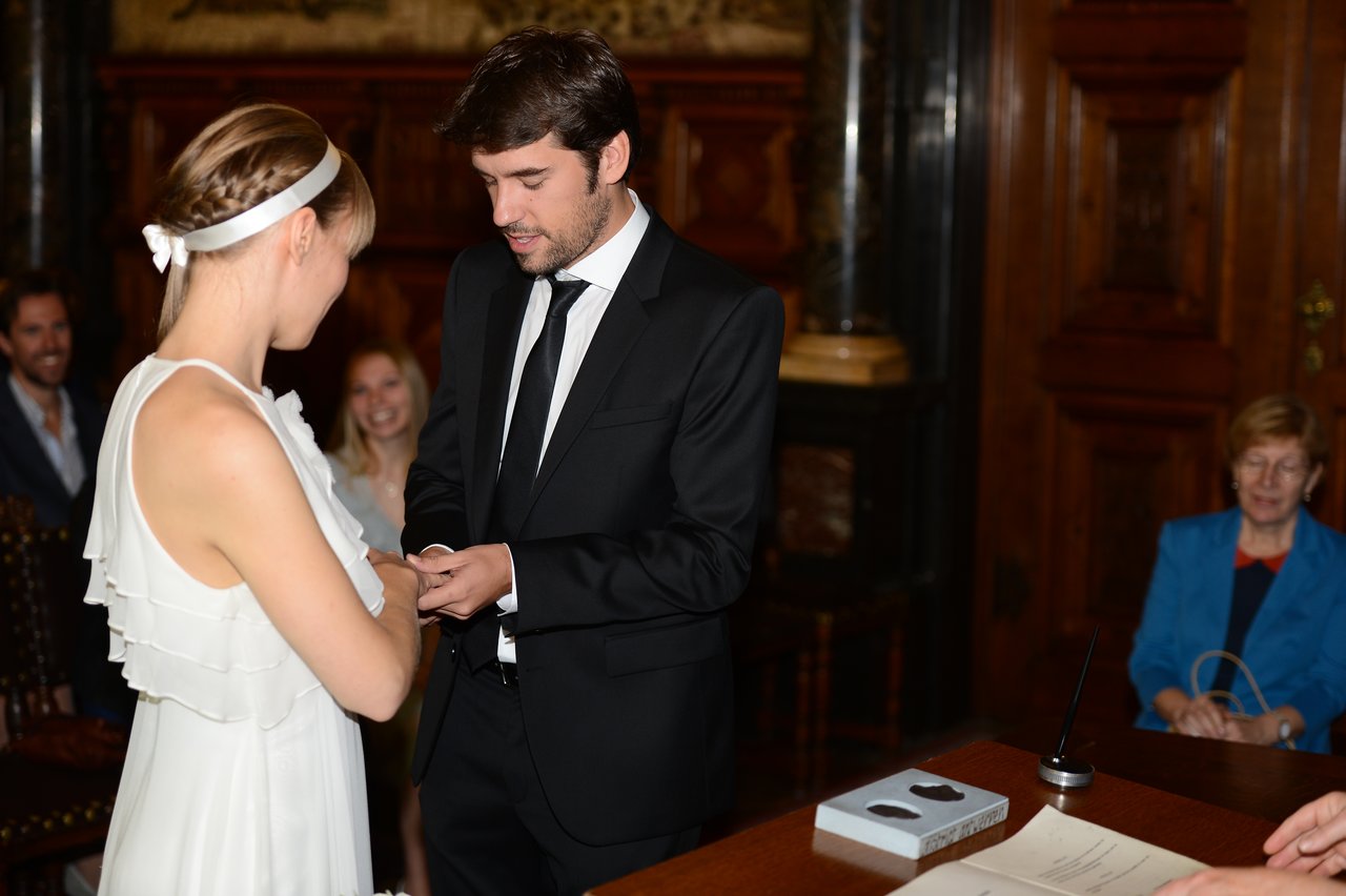 A groom places a wedding ring on the bride's finger during their marriage ceremony, with guests watching.