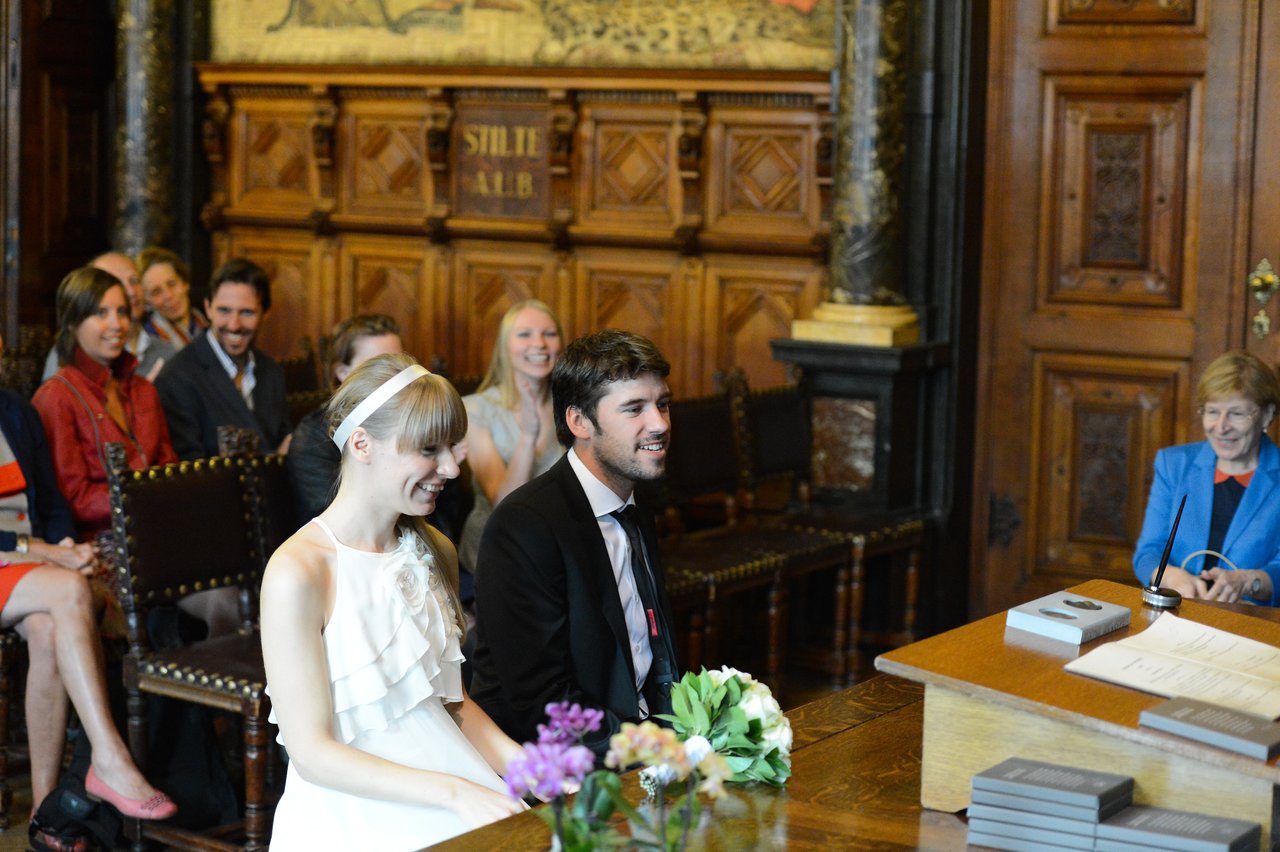 A bride and groom sit together, smiling, during their wedding ceremony in a formal room with guests watching.