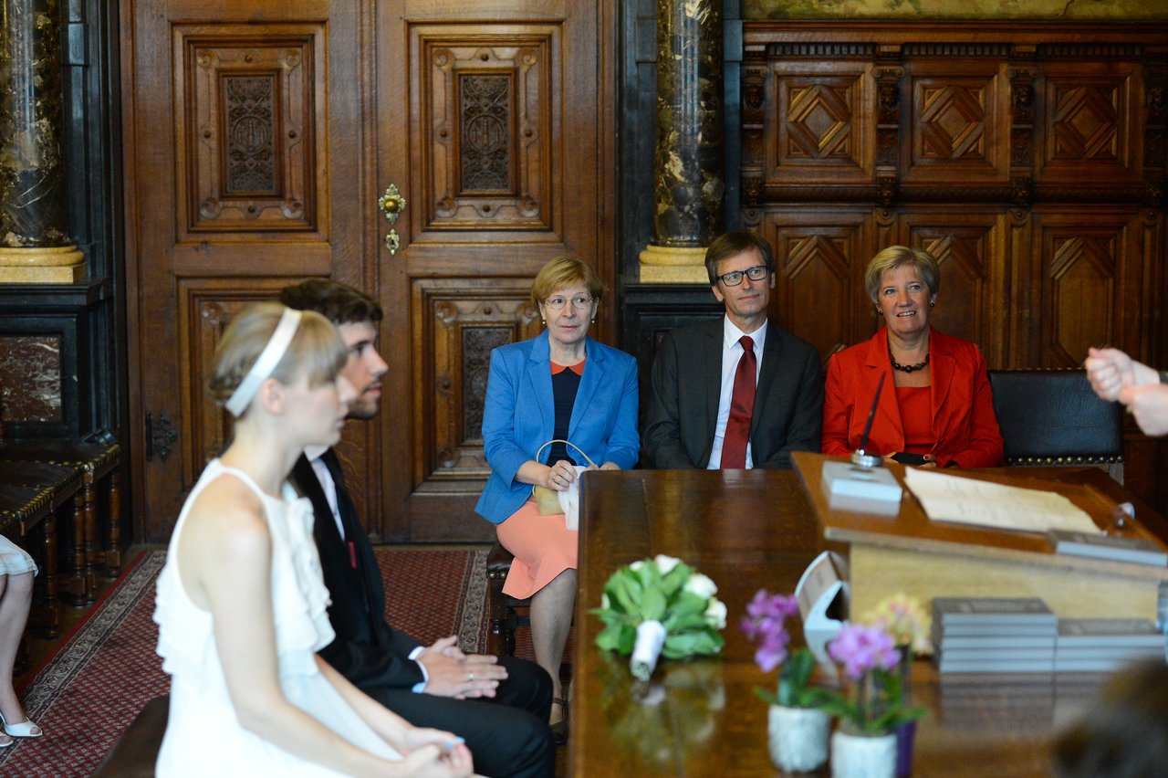 A bride and groom sit at a table during their wedding ceremony, with guests watching from behind.