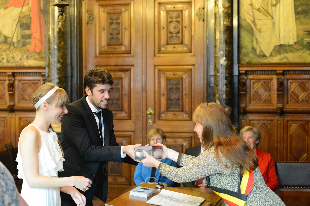 A bride and groom receive a small box from an official during their wedding ceremony in a formal setting.