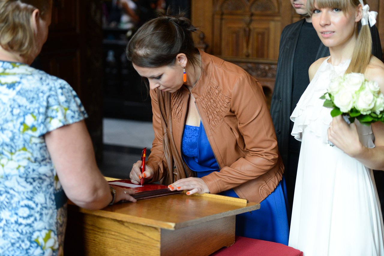 A woman in a blue dress signs a document while another woman in a white dress holds a bouquet.