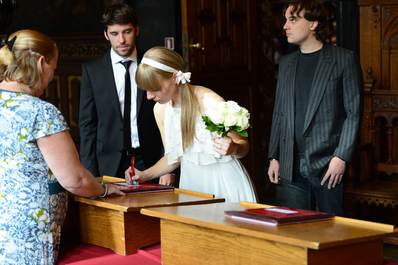 A bride in a white dress signs a document while holding flowers, with others watching in a formal setting.