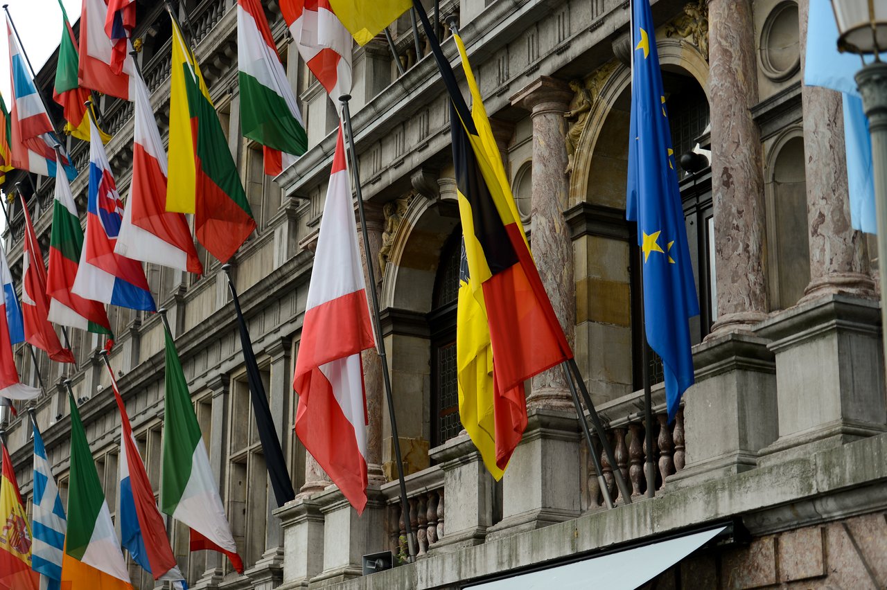 A building with many international flags hanging from poles, including the European Union and Belgian flags.