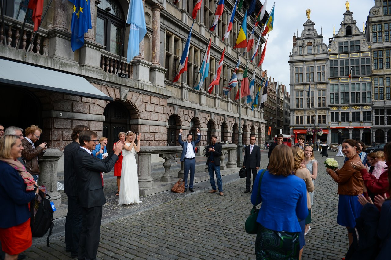 A bride in a white dress claps while guests cheer and a woman holds a bouquet in a public square.