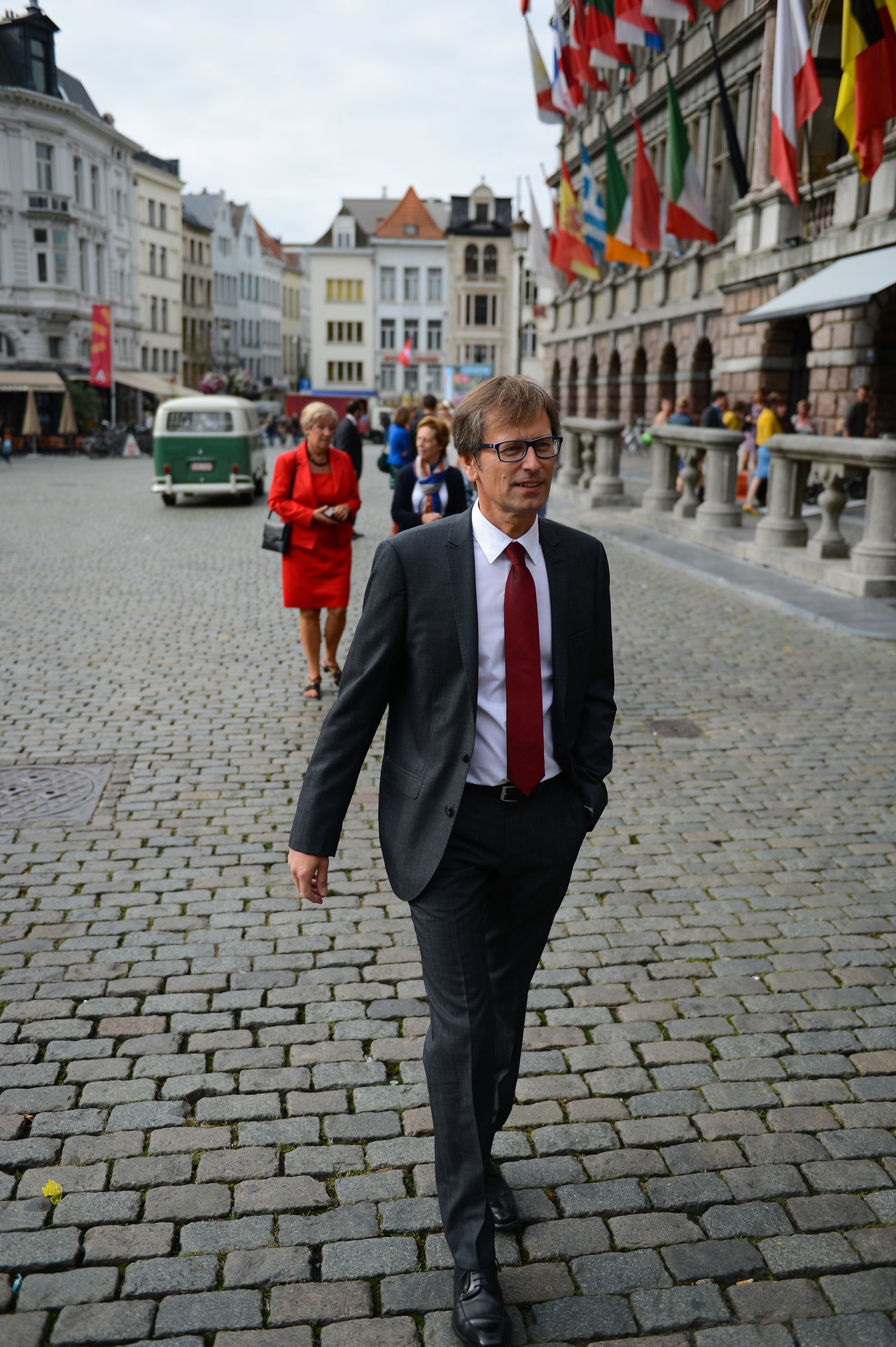 A man in a suit with a red tie walks on a cobblestone street, with guests behind him.