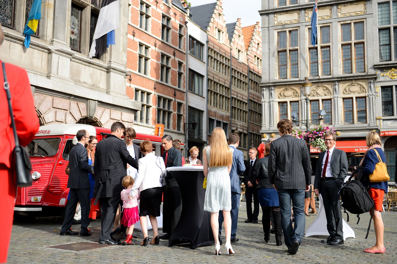 Wedding guests gather and socialize in an outdoor plaza, dressed in formal attire, with a red vintage van nearby.