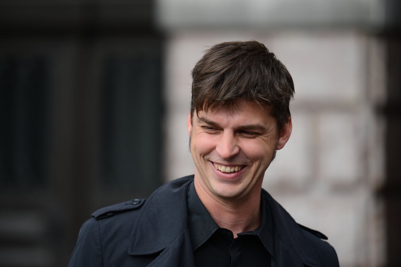 A man in a dark coat smiles while looking down, standing outside a building.