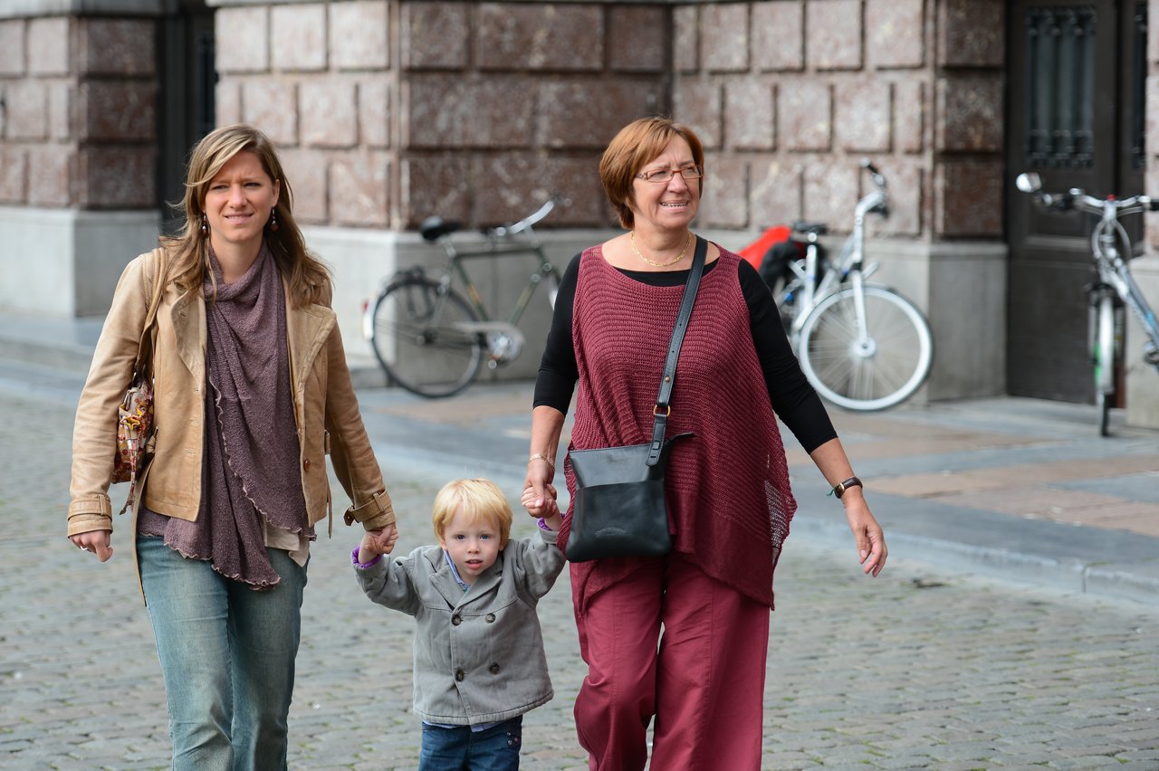 Two women walk with a young child, holding hands, on a cobblestone street near bicycles and a building.