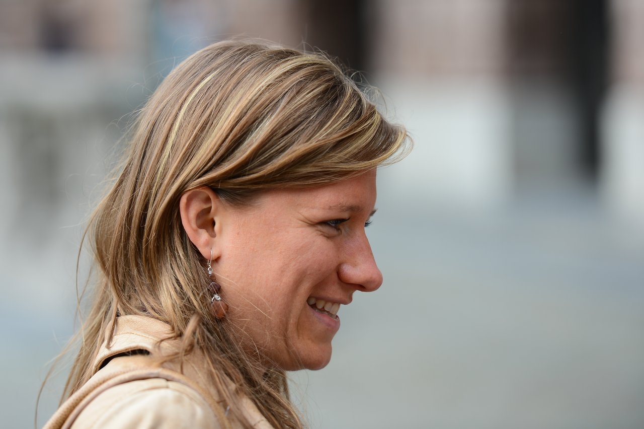 A woman with long blonde hair smiles while looking to the side, wearing a beige jacket and dangling earrings.