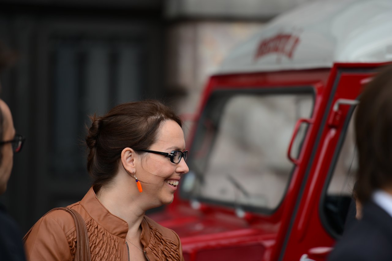 A woman in a brown jacket smiles while talking to others near a red vehicle.