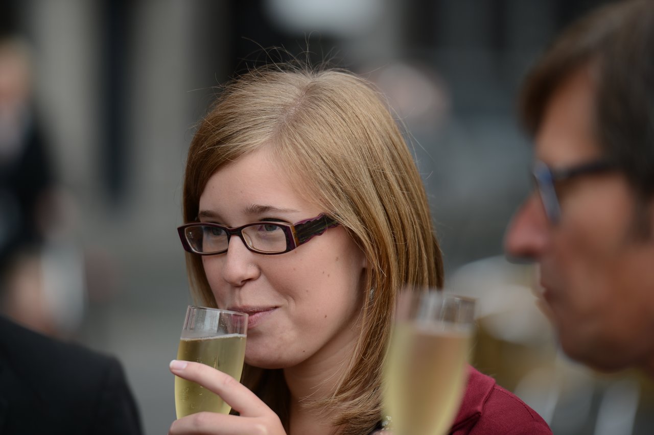 A woman wearing glasses sips champagne while attending a wedding celebration.