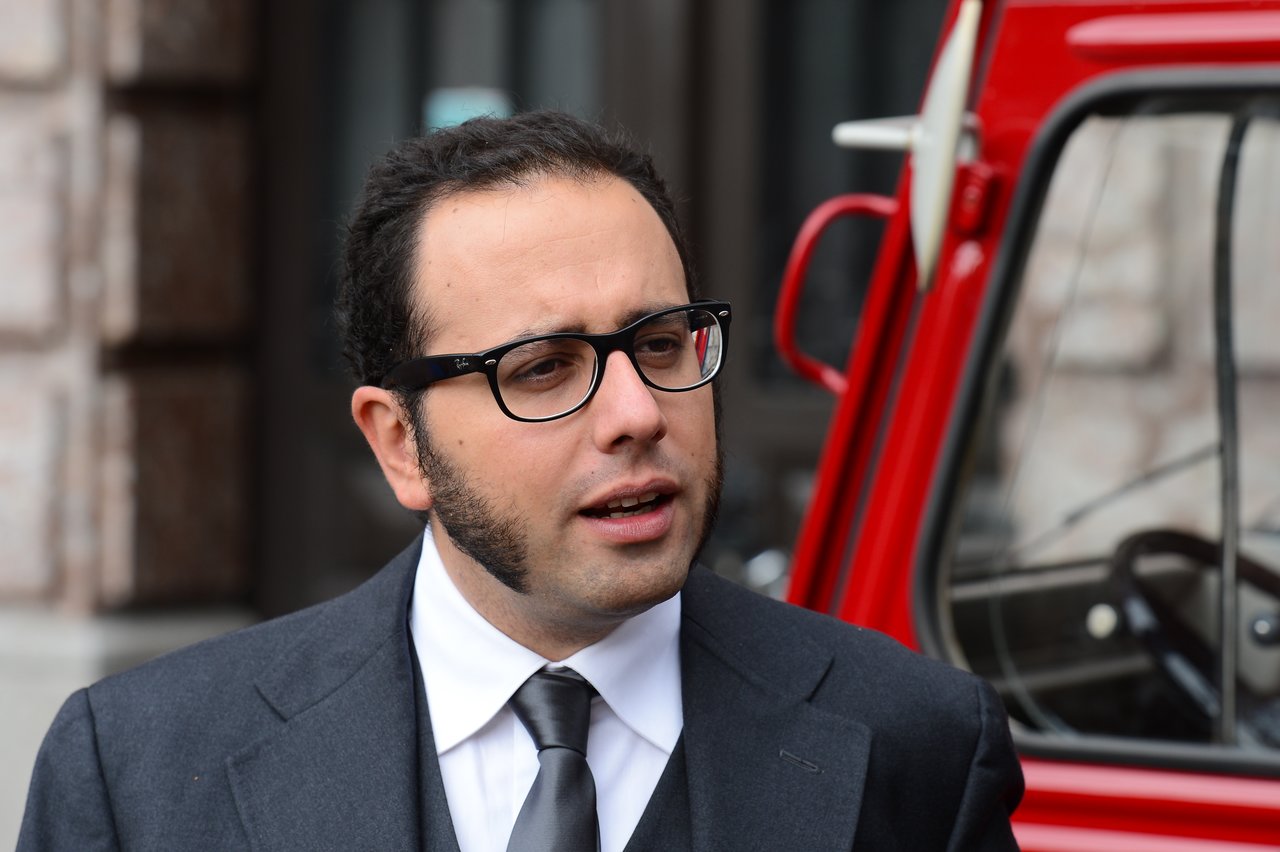 A man in a suit and glasses speaks outside near a red vehicle.