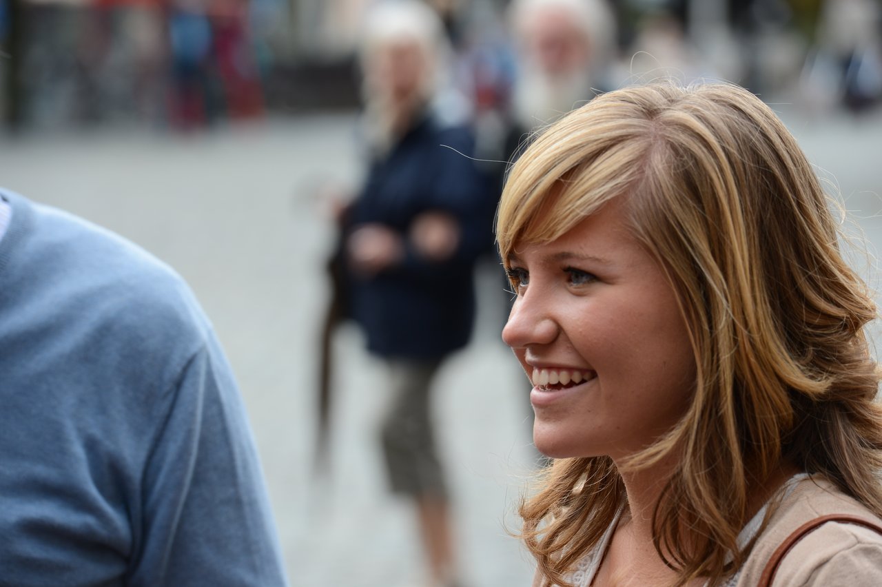 A smiling woman with blonde hair looks towards someone, with a blurred crowd in the background.
