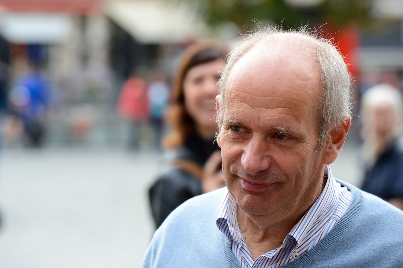 A man in a light blue sweater smiles slightly while standing outdoors, with a blurred crowd in the background.