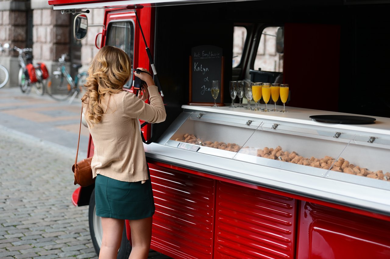 A woman takes a photo of a red food truck with a display of drinks and snacks.