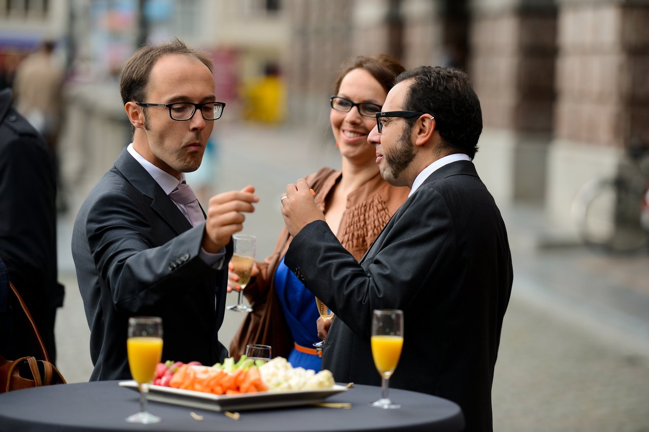Three wedding guests in formal attire stand by a table, chatting and enjoying drinks and appetizers.