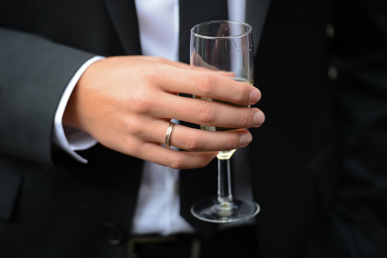 A person wearing a wedding ring holds a champagne glass, dressed in a dark suit.