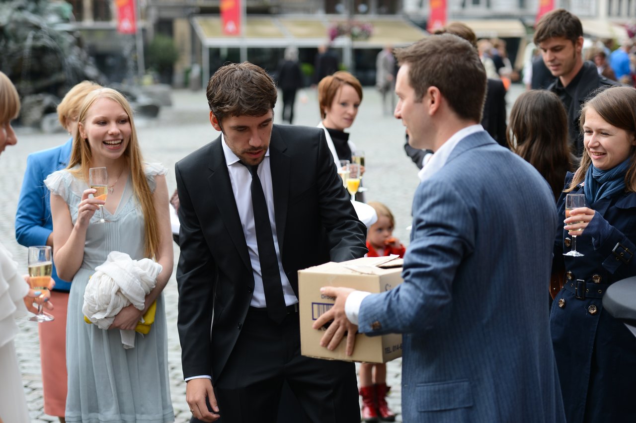 A man in a suit hands a cardboard box to another man at an outdoor wedding gathering.