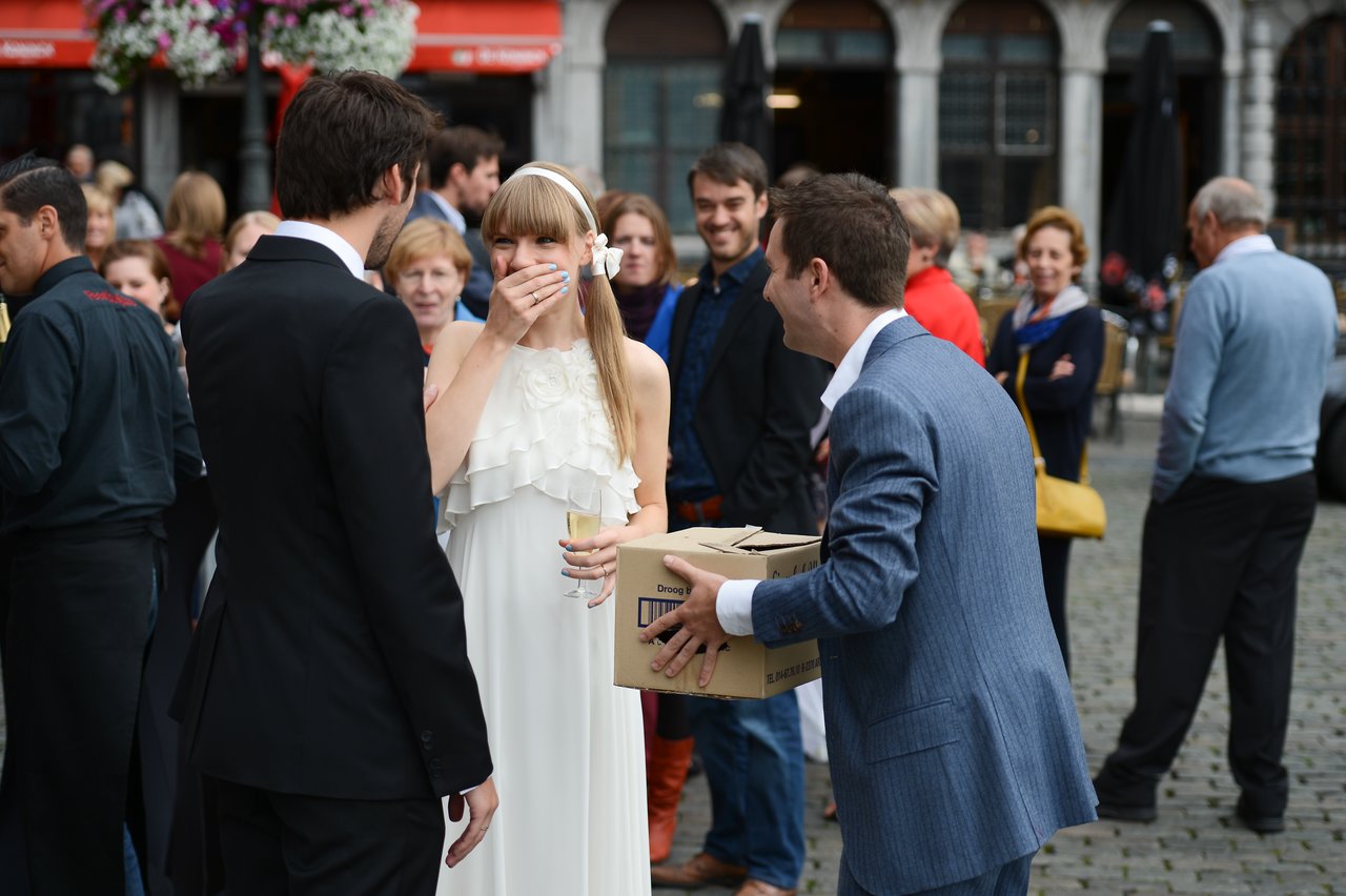 A bride in a white dress laughs while holding a drink, as a man in a suit hands over a box.