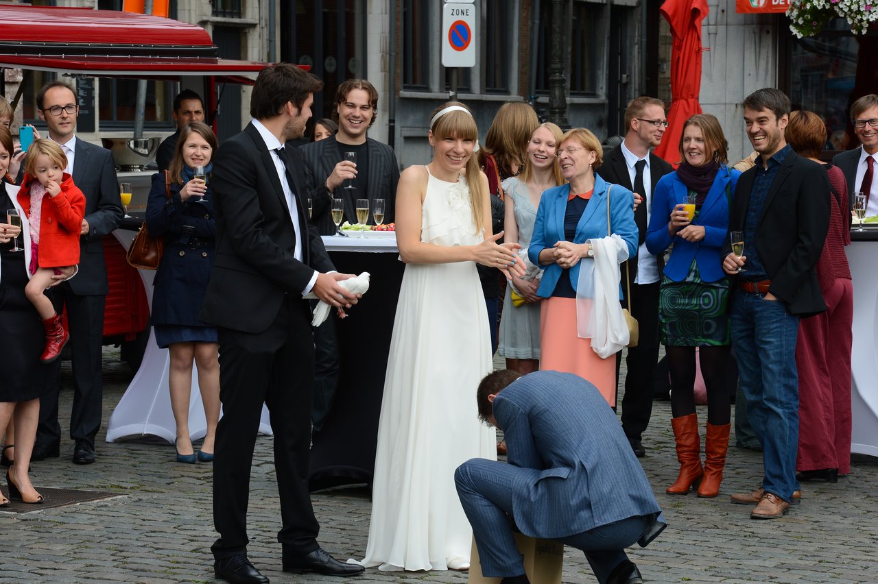 A bride and groom laugh as a man kneels in front of them, surrounded by wedding guests.