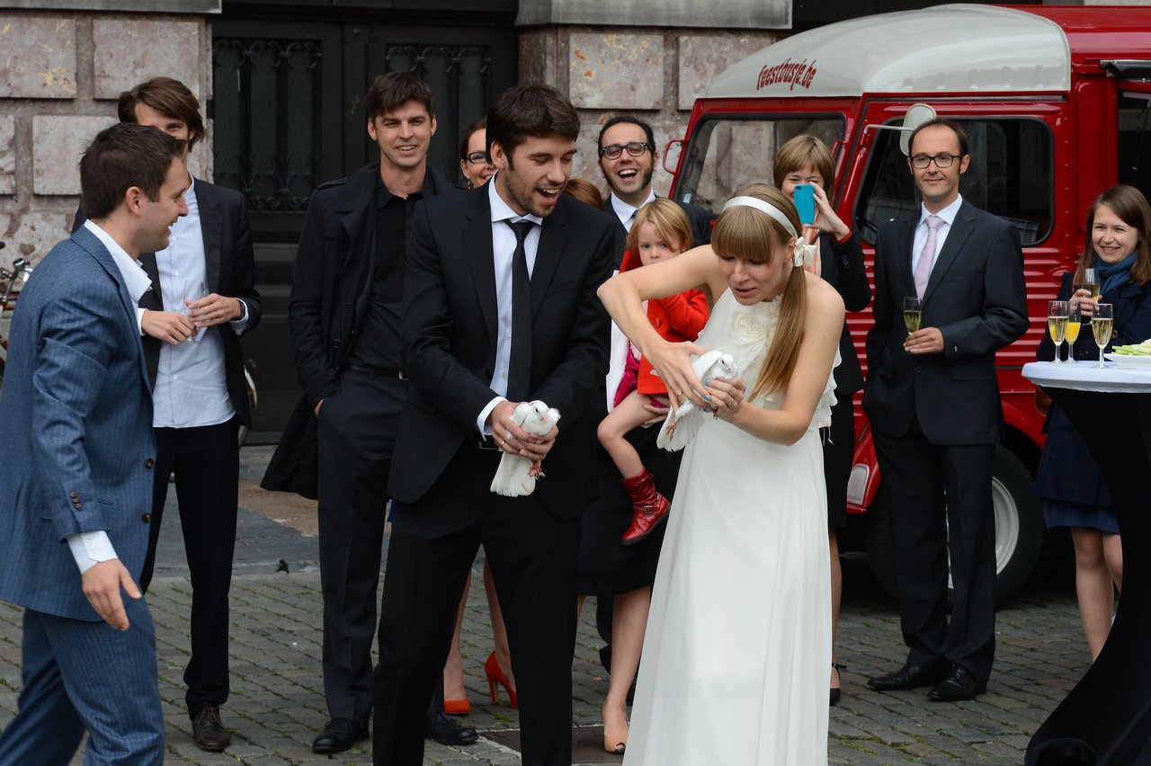 A bride and groom hold white doves while laughing, surrounded by wedding guests in formal attire.