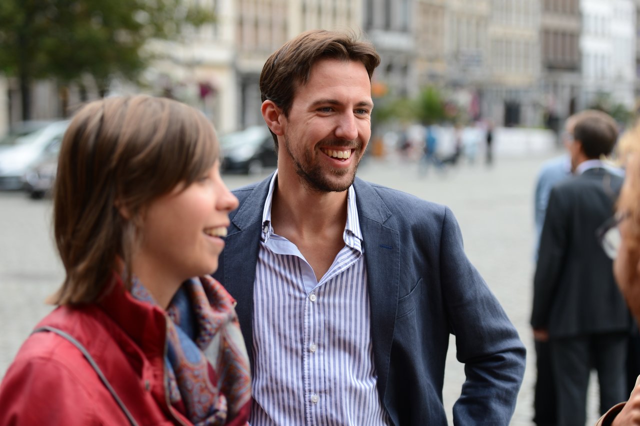 A man in a suit smiles while talking with others at an outdoor wedding gathering.