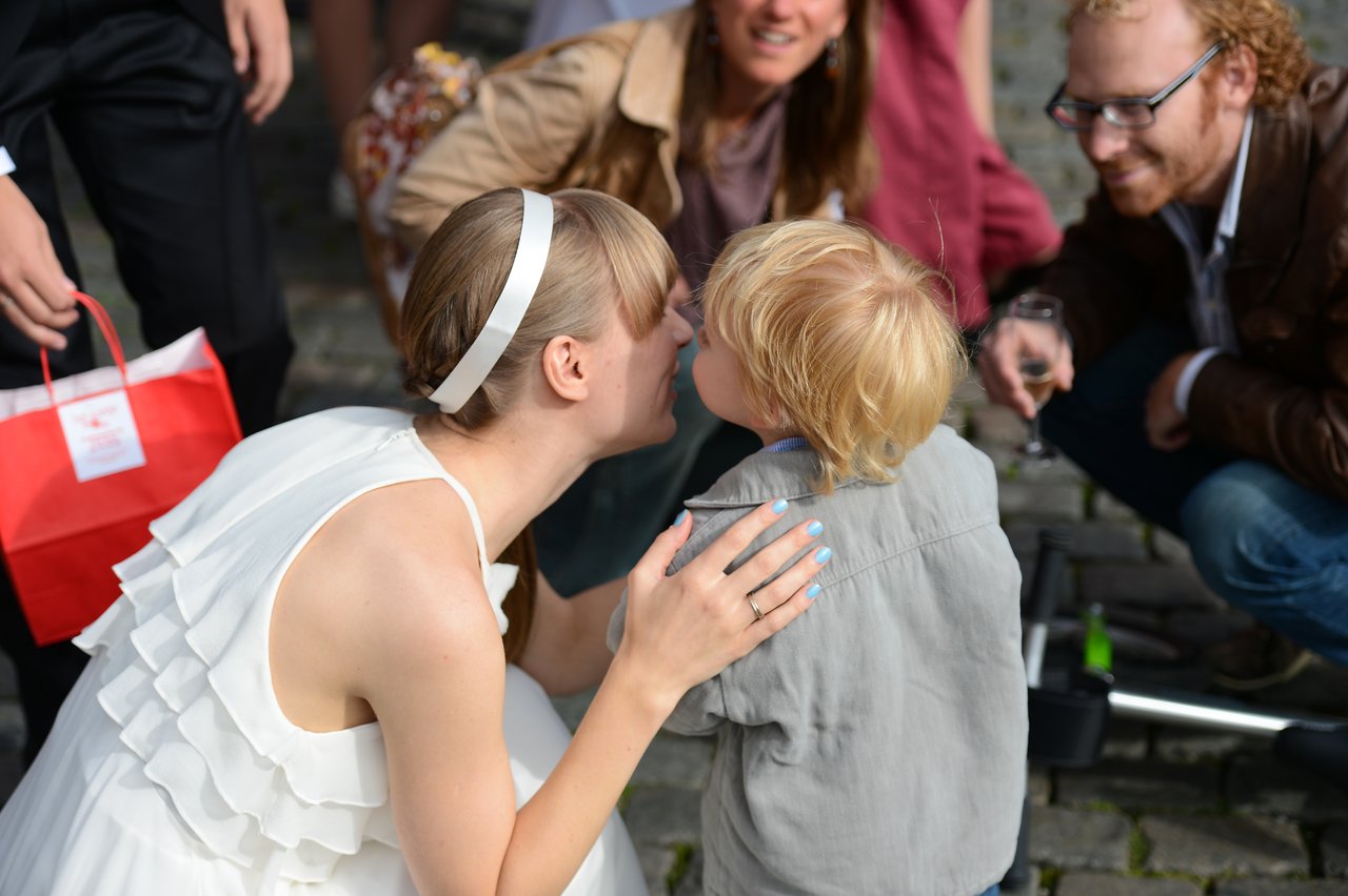 A bride in a white dress leans down to kiss a young child, surrounded by smiling guests.