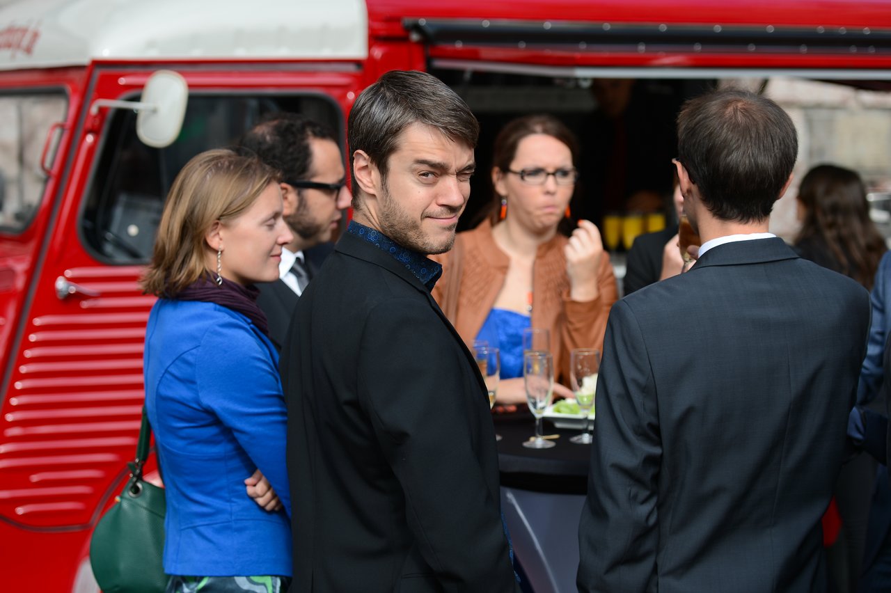 Guests in formal attire stand and chat at an outdoor wedding reception, holding drinks near a red food truck.