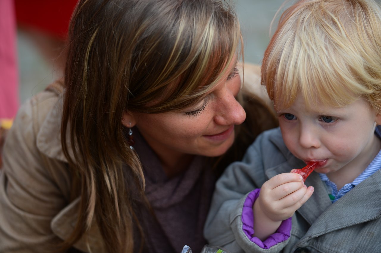 A woman smiles while looking at a young child eating a red candy.