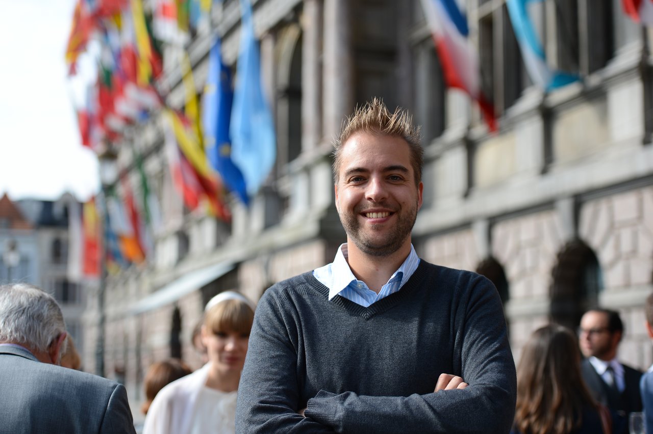 A smiling man stands with arms crossed, while wedding guests gather in the background near a building with flags.