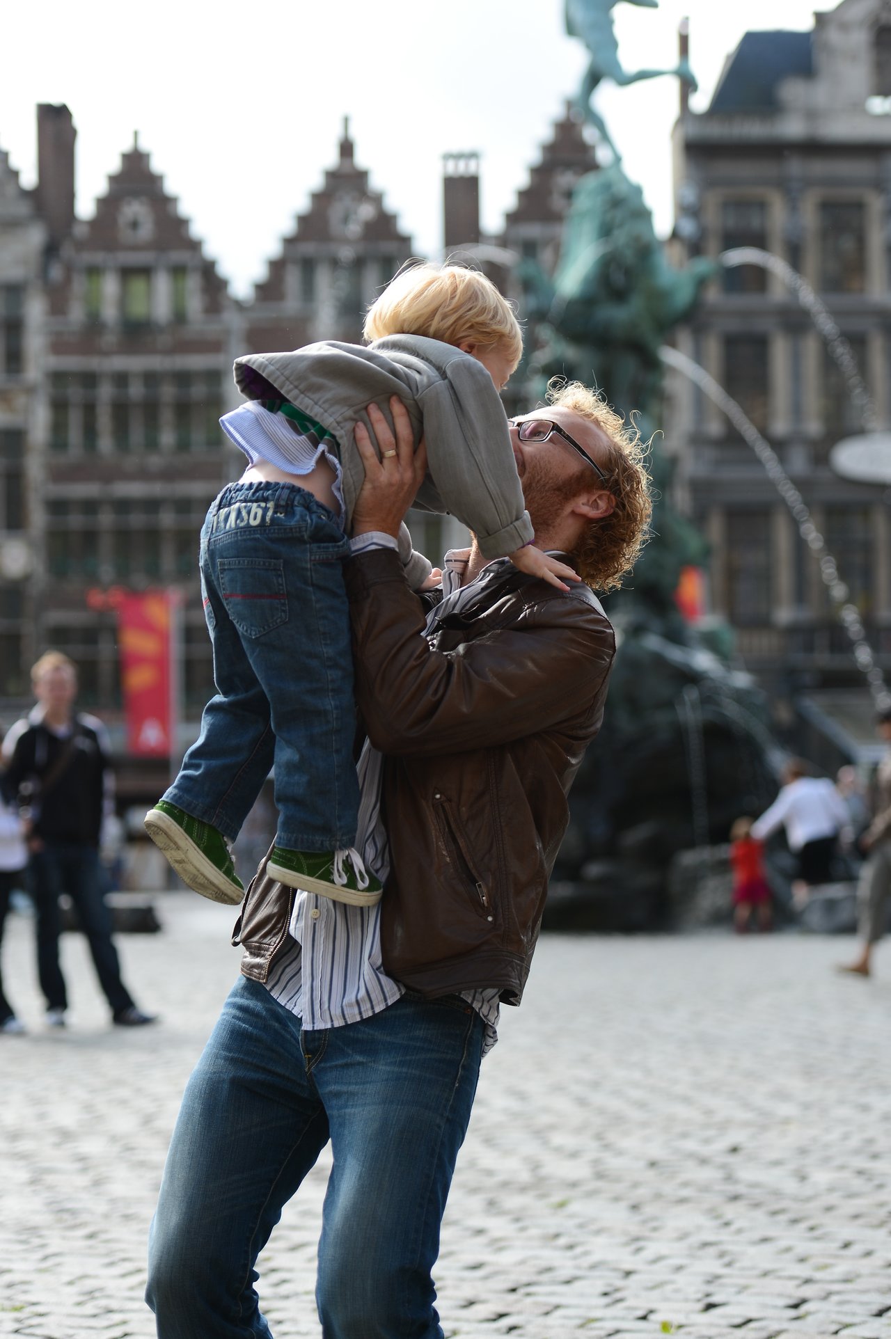 A man lifts a young child in the air while smiling in a public square with historic buildings.