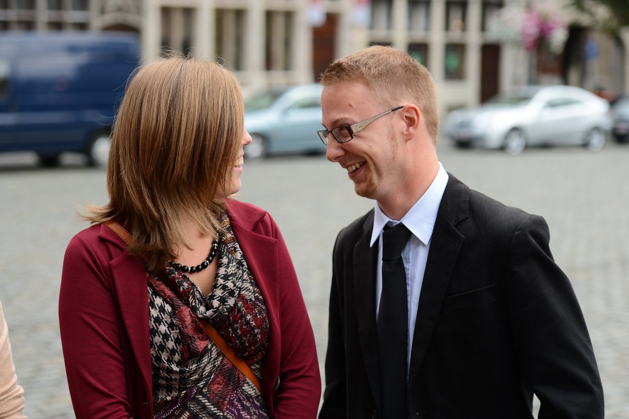 A man in a suit and a woman in a red jacket smile at each other during a wedding event.